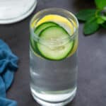 Cucumber Water in a glass placed on a grey table with lemon and mint leaves around